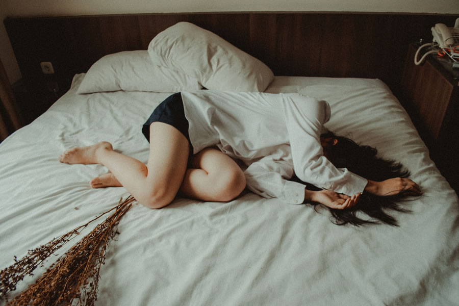 a woman in a white blouse and black shorts laying on a bed with white sheets covering her face