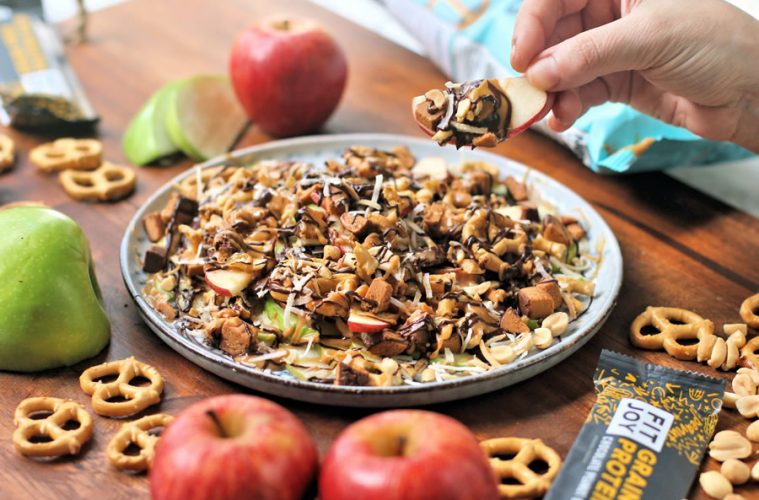 a hand holding up an apple slice with chocolate sauce peanut butter coconut peanuts and FitJoy protein bar crumble